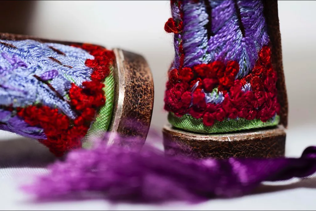 A pair of vibrantly coloured medium Louis heeled shoes decorated with an array of feathers and embroidery stitches including hundreds of French knots. The shoes feature a predominantly green and red pattern, accented with splashes of purple, pink, and blue. A purple satin ribbon ties at the instep. Here we can see the heels with highly detailed embroidery work covering them in a dream like vision.