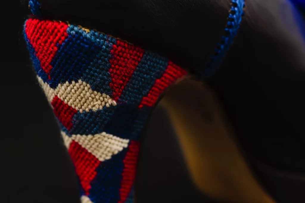 Zoomed in detail of the heel of the Campaign shoe by Caroline Groves. Here you can see the heel is covered in needle point work of a geometric design. This embroidery is in a deep blue, red and white. The dark grey of the veg tanned leather upper is just visable and a rich blue hand stitched detail runs up the edge of the shoe.