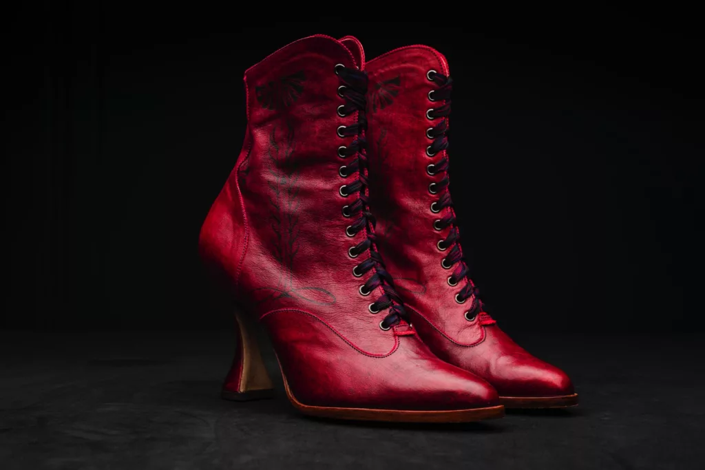 This photo shows a pair of deep red Caroline Groves leather booties against a black background, accentuating their exquisite, dramatic style. The heel is a fairly high and with a curvaceous top piece, the ankle boot is a slim fit at the ankles and has an almond toe shape. These ankle booties have 14 eyelets and a grosgrain lacing of deep purple and magenta hues. A hand inked decorative art piece runs up the quarter panels, inspired by the binding of a Keat’s poetry book.