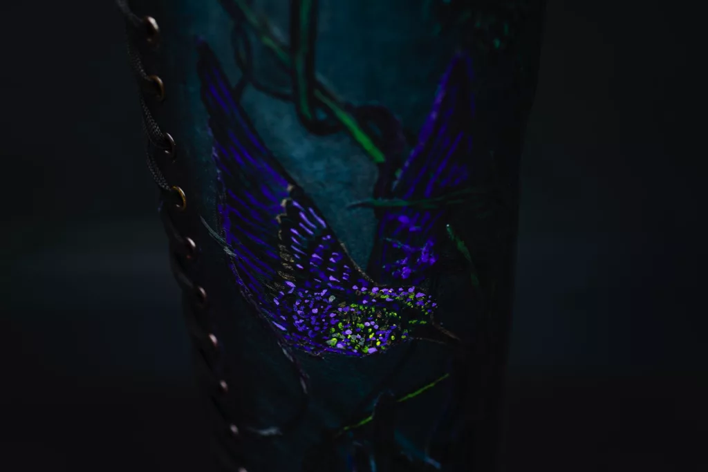 A close up of a full length, deep blue lace up boot. Showing a bird that has been intricately inked and painted onto the leather . The boots dark metal eyelets are just shown in the photo with a black silk lace running through them.