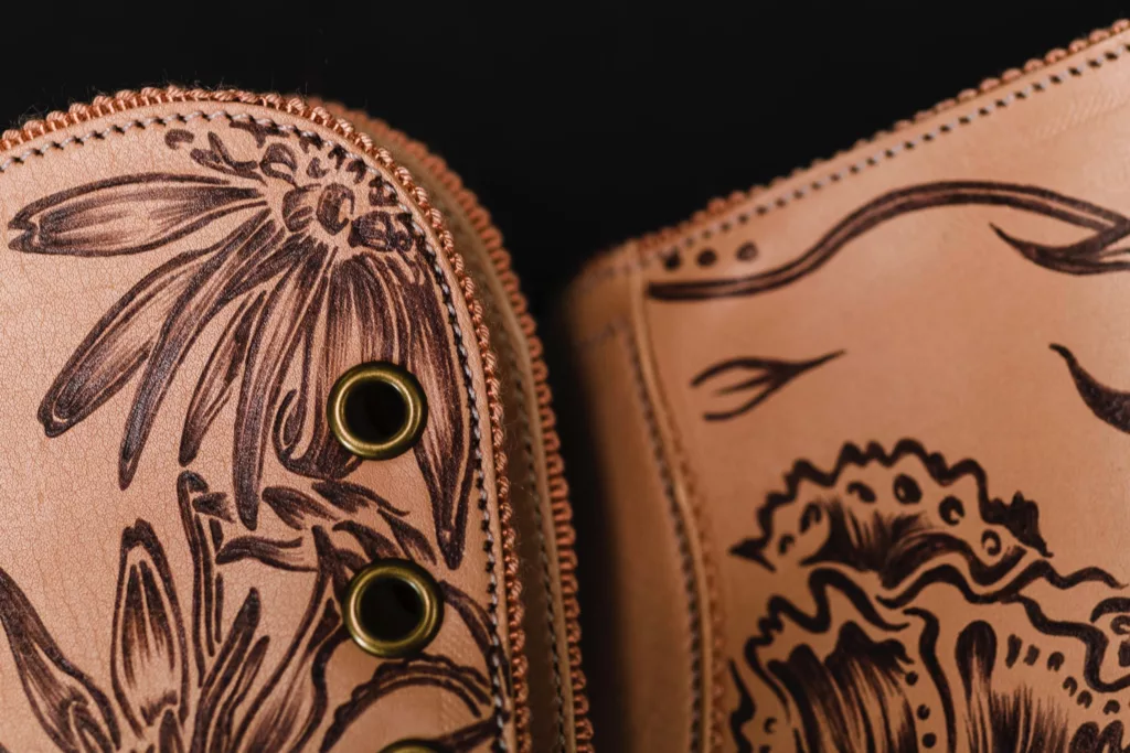 Close up of a pair of boots in a harmonious colour palette, with brass metal eyelets , nude complimentary grosgrain beading at edges. The main body being a nude leather of vegetable tan, with sepia art work representing the flowers of Kazakhstan hand inked directly onto the leather freely but with highly refined.