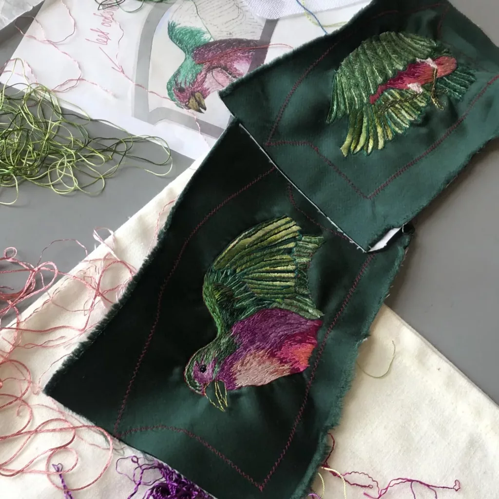 Embroidery In Progress - Caroline Groves. Here you see the satin dark green heel coverings layer out on a table with the embroidery of the exotic bird well under way. Bright and glistening the silks and gold threads look fantastic .