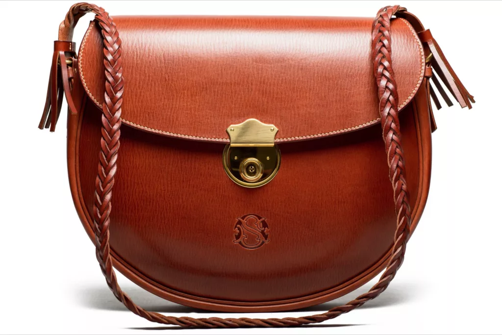 This photo shows the ‘ Cadley’ shoulder bag by Caroline Groves, featuring the deboss detail on its vegetable tanned leather, a rich tan colour, the deboss being rounded in shape and depicting the clients own monogram, exquisitely pressed into the leather. The brass clasp is above the deboss and of a high shine. The bags plaited leather strap hangs in front of it and it has matching tassles fixed to each end. The bags edges are hand stitched to perfection.