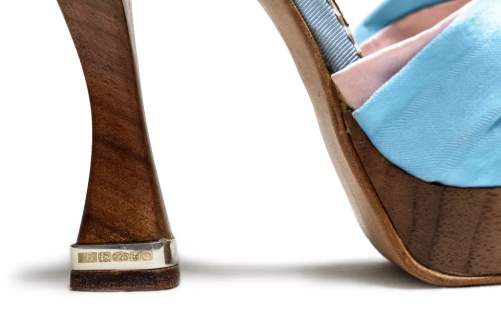 A pair of impactful statement platform sandals. Using the wings of the turquoise parakeet bird to decorate the back counters and solid silver at the front in the form of a striking claw. The claw sits on an upper of blue vintage French silk. A solid Silver heel band and solid walnut is the chosen material for its platform and heel, which you can see here a close up of, included the hallmarks on the silver heel band.