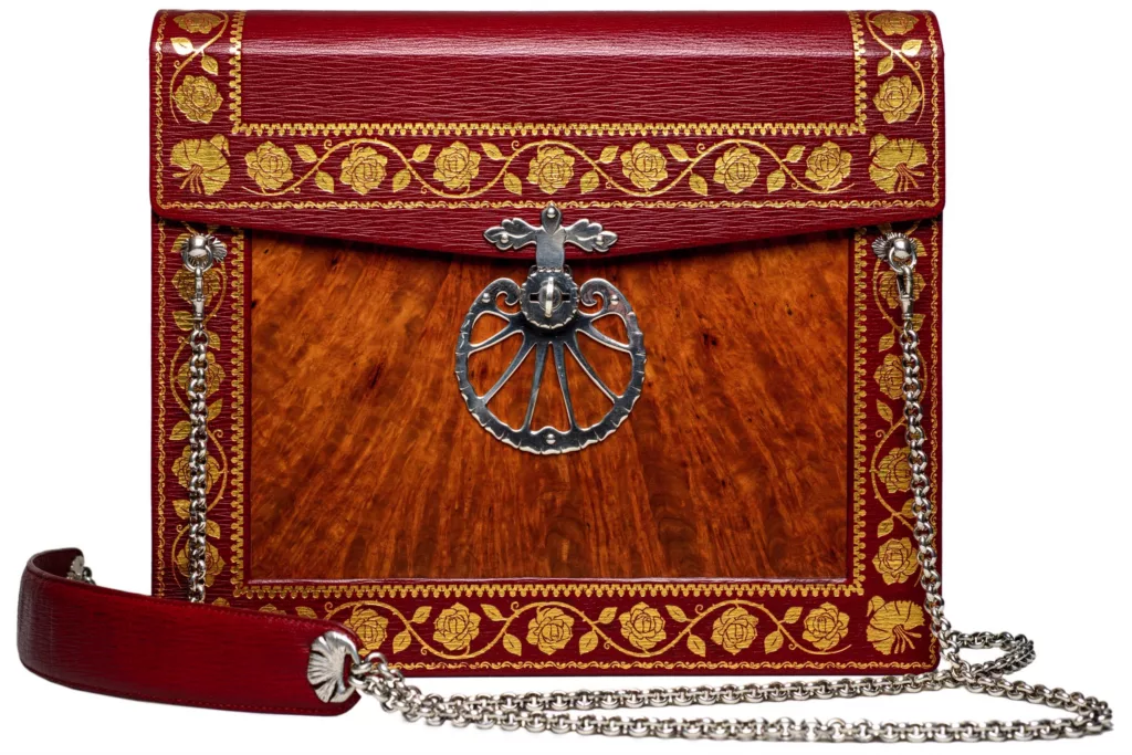 Photo revealing the detail of the front of the ‘Dovers Reticule’ Shoulder case, inspired by an English period reticule. Red goat embossed and tooled with roses and pinks. Inlayed with Burr-Amboyana Wood Veneer. Solid Silver two part swivel mechanism clasp with engraving and patterned edges. ‘Fish tail’ handle. Fully hand stitched with linen thread. The metal chain of the shoulder strap hangs from two special attachments and has a leather section which sits on the wearers shoulder with comfort.