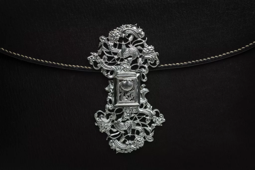 This photo shows the close up of a highly detailed solid silver clasp, craftsman made, sitting as center piece on this elaborate side of the Smerrel design. The clasp depicting magical bird details, florals and shell-like shapes. This side of the bag is in black vegetable tanned leather with an ornate embossing floral detail on main body and following the curve of the flap. Perfectly executed hand stitching follows the edges of the leather.