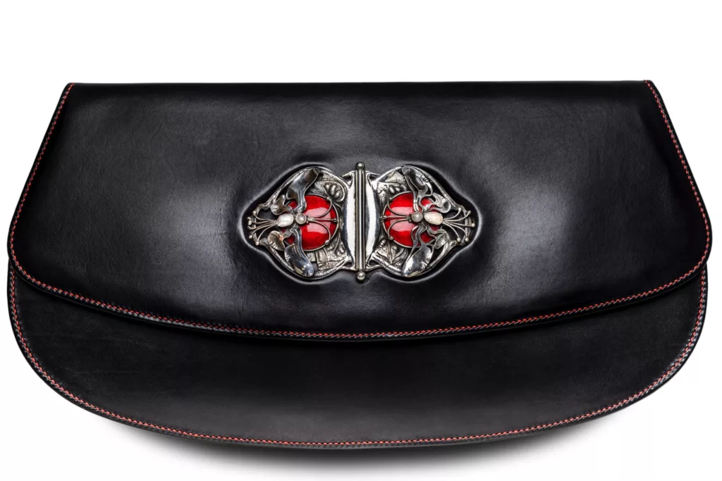 Close up of black vegetable tanned leather clutch bag. Fine red stitching detail and incredible solid Silver Antique Buckle with red enamel. Buckle is of the Ashbee’s guild handcraft, depicting a mirror image of a set of dragonflies wonderfully stylised from the early 19 hundreds. Buckle is set into a moulded cavity at center front of this elegant clutch bag.