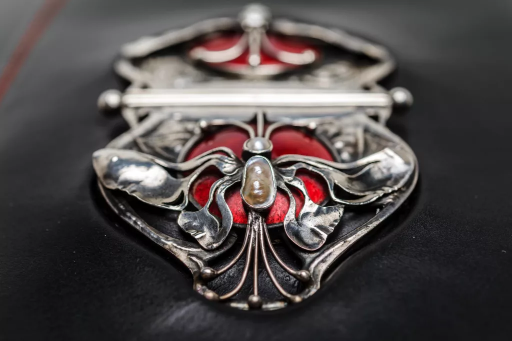 This photograph is a close up of part of an antique buckle carefully moulded into a Caroline Groves black vegetable tanned clutch bag. The buckle is solid silver and intricate with floating wings and further stylised metal work decoration of a circle of red enamel.