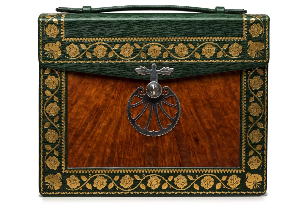 Photo revealing the detail of the front of the ‘Dovers Reticule’ Shoulder case, inspired by an English period reticule. Green goat embossed and tooled with roses and pinks. Inlayed with Burr-Amboyana Wood Veneer. Solid Silver two part swivel mechanism clasp with engraving and patterned edges. ‘Fish tail’ handle. Fully hand stitched with linen thread. The metal chain of the shoulder strap hangs from two special attachments and has a leather section which sits on the wearers shoulder with comfort.