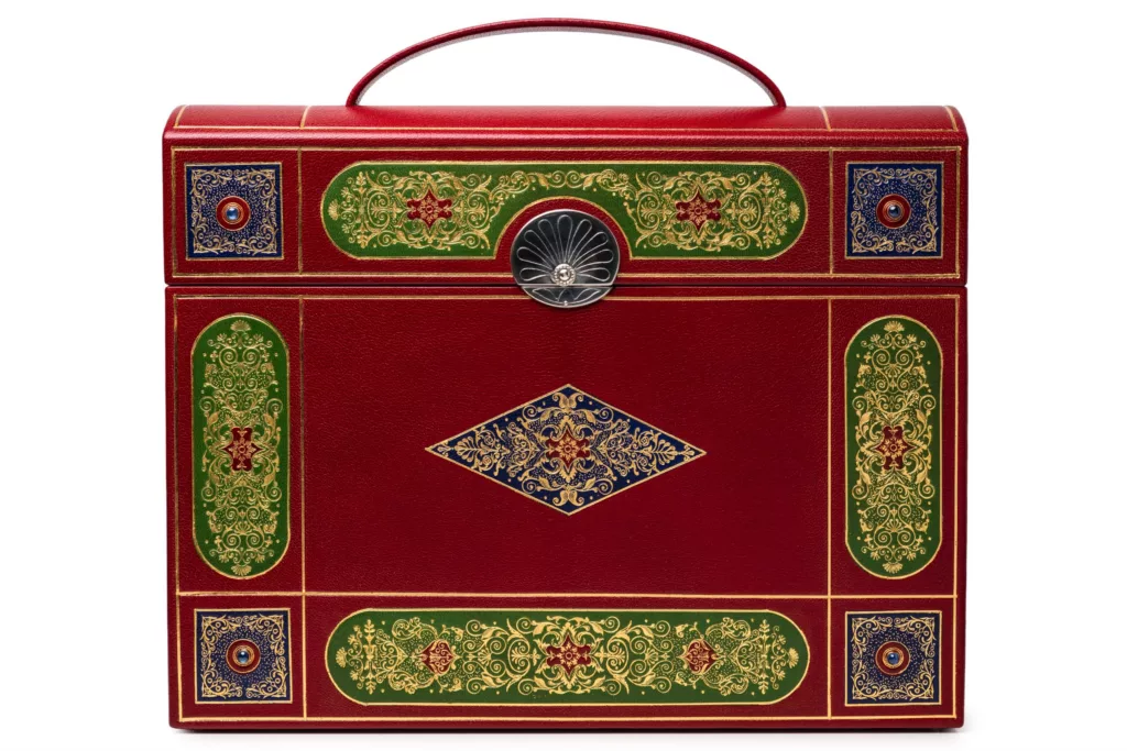 A photo of a highly detailed case which appears like a bound book of great importance, only with a top handle revealing itself as a carry case of high sophistication. The Cleeve Case for Caroline Groves is inspired by late Georgian period artifacts. In red goat skin with onlays of green and blue. It is a heavily tooled piece with 22t gold. At the corners, sapphires are set in gold bezels. With a solid silver engraved slide clasp, silver hinges, screw caps and domed pin feet. Lined in green gold with simple hand-sewn pockets.