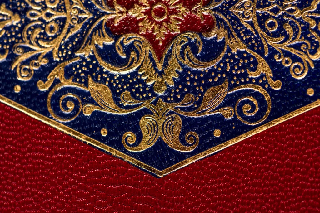 A close up photo of part of a highly detailed case, showing the 22t gold tooling on a dark blue goat leather and red goat leather section. The Cleeve Case for Caroline Groves is inspired by late Georgian period artifacts. In red goat skin with onlays of green and blue. It is a heavily tooled piece with 22t gold. At the corners, sapphires are set in gold bezels. With a solid silver engraved slide clasp, silver hinges, screw caps and domed pin feet. Lined in green gold with simple hand-sewn pockets.