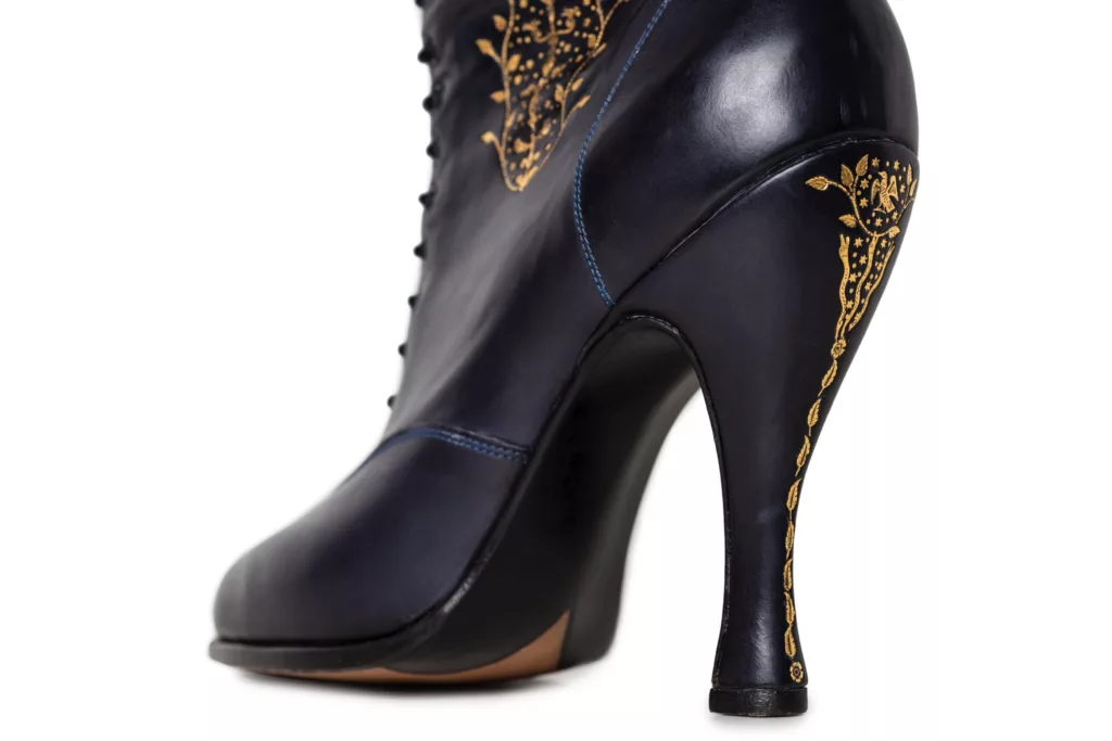 The back view of this deep blue book calf leather boot. Seeing the carefully positioned 22t gold hand tooling running up the back of the heel to a detail filled finale at its top. Also within viewing range is the beginning of yet more of this intricate tooling beginning to descend up the leg panel. The imagery is inspired by temptation in the garden of Eden. This Oxford boot is long leg with laces running up all the way. This is a hand welted and hand stitched construction.