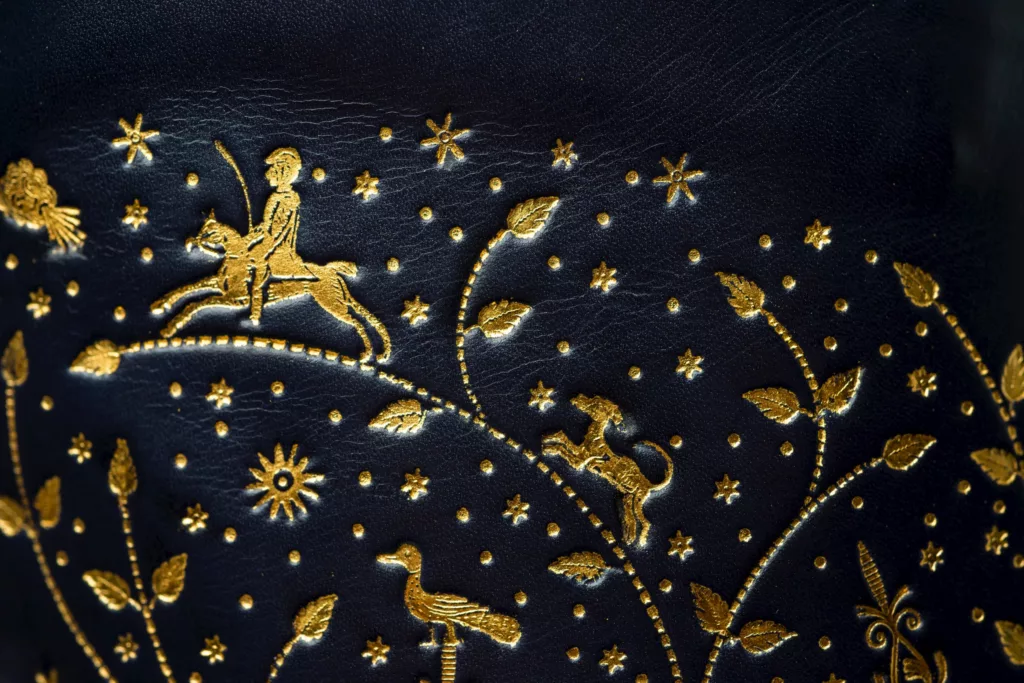 A close up of this deep blue book calf leather boot. Seeing just a small section of the carefully positioned 22t gold hand tooling detail close up. Here are leaves, stems, birds, planets and stars to name a few of the shapes. This intricate gold tooling in fact fills its leg panels and the back of a high heel. The imagery is inspired by temptation in the garden of Eden. This Oxford boot is long leg with laces running up all the way. This is a hand welted and hand stitched construction.