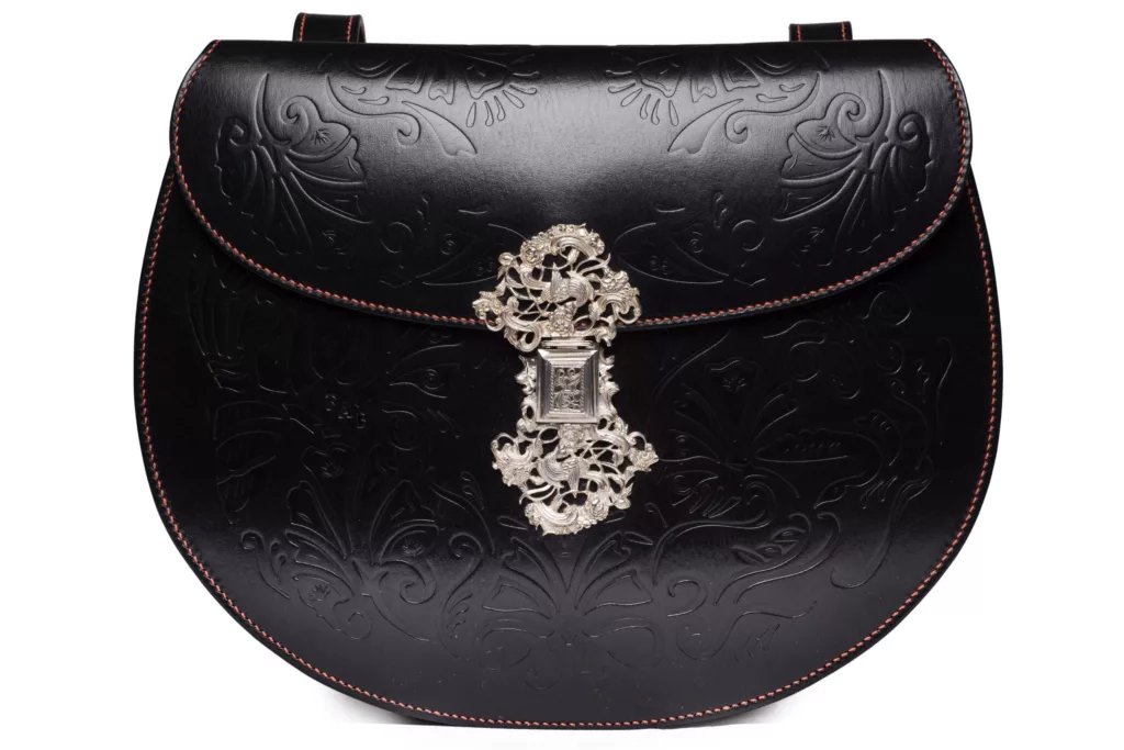 A photo of the highly detailed side of a double faced handbag by Caroline Groves. This side of the bag is in black vegetable tanned leather with an ornate embossing floral detail on main body and following the curve of the flap. Perfectly executed hand stitching follows the edges of the leather. A highly detailed solid silver clasp, craftsman made, sits as center piece on this elaborate side of the Smerrel design.