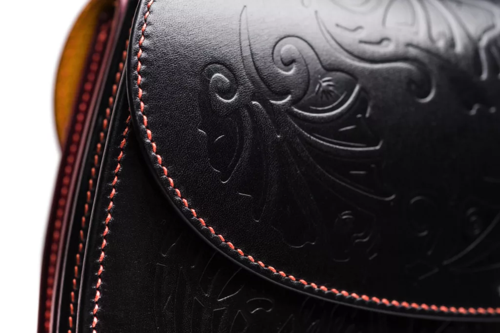 This photo shows the close up of one side of the Smerrel bag. In black vegetable tanned leather with an ornate embossing floral detail on its main body and following the curve of the flap. Perfectly executed hand stitching follows the edges of the leather in a striking red linen thread.