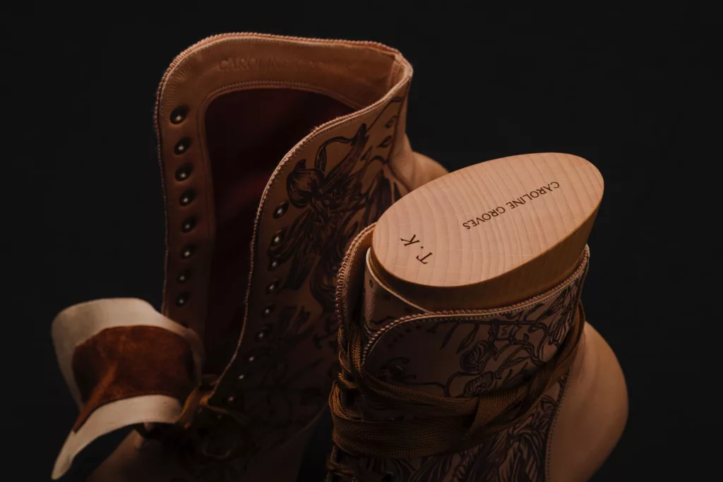 Close up of a pair of boots with perfectly carved boot trees within. A tight fitted, attractive ankle shape with laces wrapped carefully around ankle. A nude leather of vegetable tan, with sepia art work representing the flowers of Kazakhstan. A complimentary sepia grosgrain lace passes loosely through the metal eyelet holes. The boot tree has the stamp marks of both their maker and initials of the client who owns them.