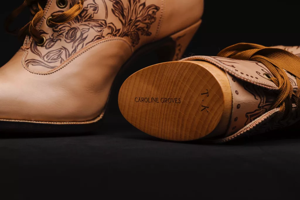 Close up of a pair of boots with perfectly carved boot trees within. A nude leather of vegetable tan, with sepia art work representing the flowers of Kazakhstan. A complimentary sepia grosgrain lace passes loosely through the metal eyelet holes. The boot tree has the stamp marks of both their maker and initials of the client who owns them.