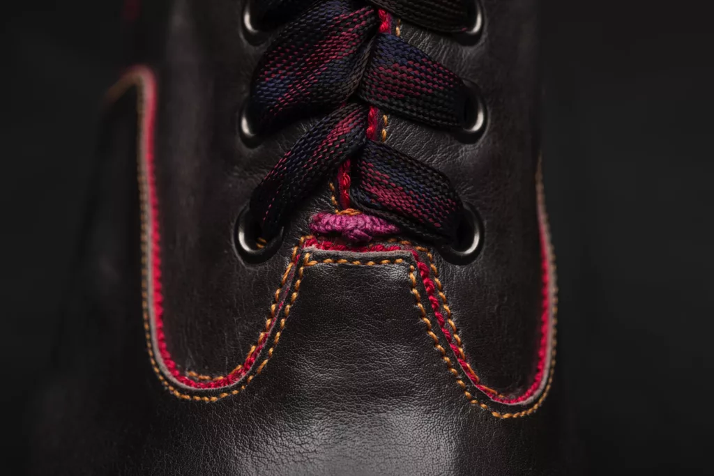 A close up front view of an Oxford bootie by Caroline groves. On black leather you can see the detail of contrasting delicate amber stitching, fiery grosgrain beading at edges and magenta hand stay stitch. Dark metal eyelets with woven magenta and other hued laces.