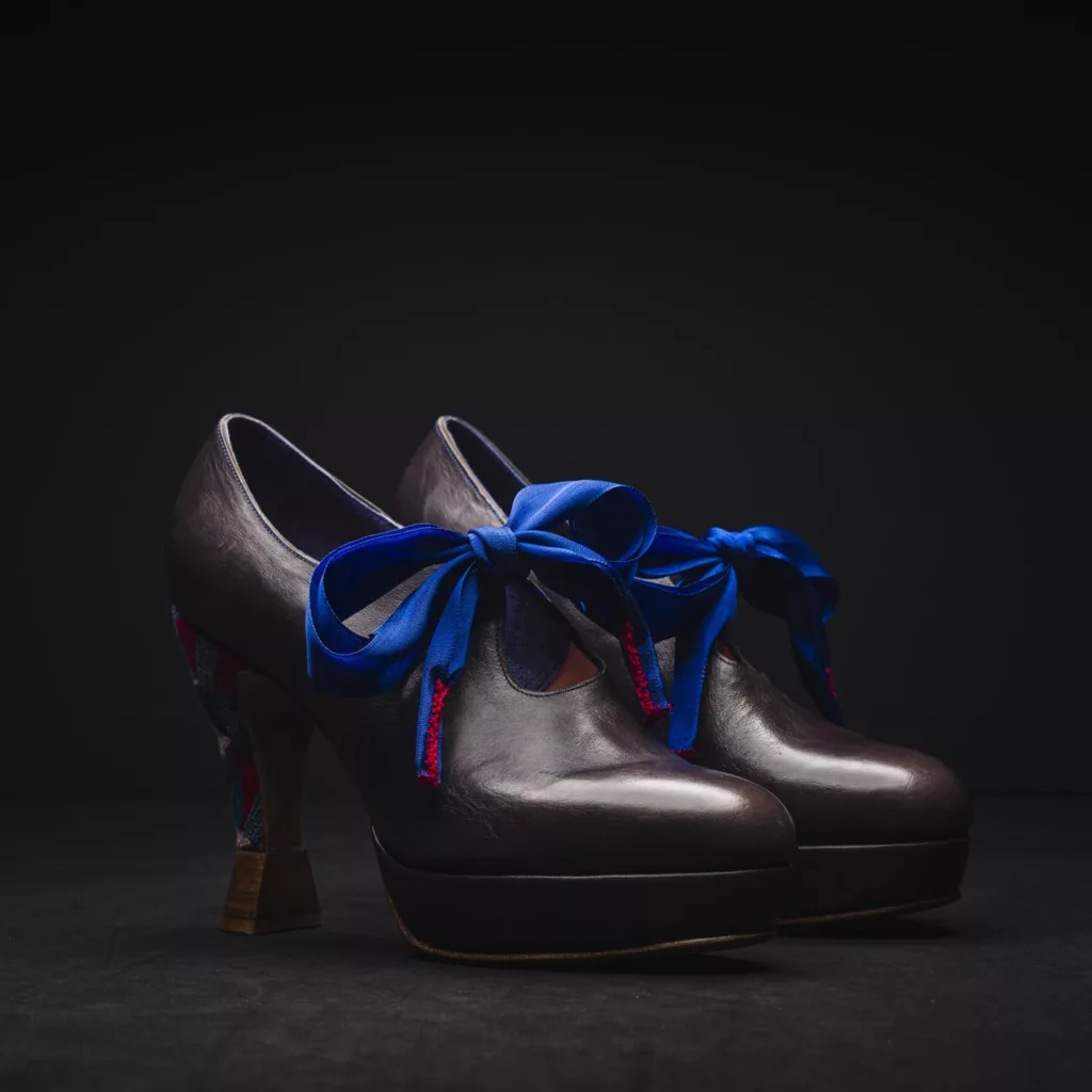 Campaign The Campaign high vamp court shoe is folk inspired. with a Louis heel covered in needle point embroidery and with multiple leather stacks. The laces are a Vintage silk French ribbon in a striking blue. Hand stitched details of this same blue are featured throughout the upper, which is a grey Veg tanned leather.