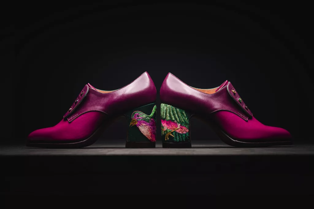 Side view of a pair of button shoes in vibrant magenta polished calf leather. With 3 Button fastenings on each foot, the main detail of this pair is upon its heel. The block heel showcase a fine work of silk embroidery, depicting an exotic bird in flight. The bright rich colours of the silk embroidery make this an exception piece of art. The shoe has a hand sewn welt and hand stitched oak bark tanned sole.