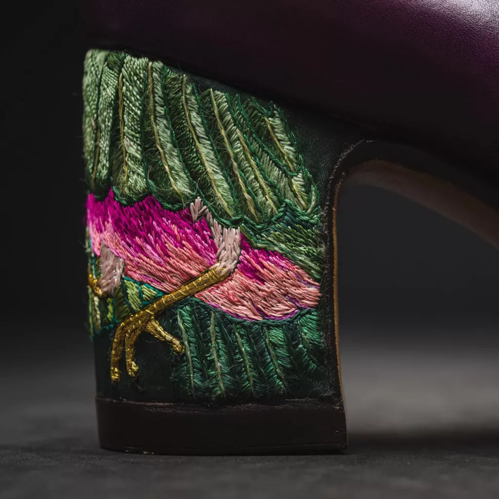 Close up heel shot of a button shoe in vibrant magenta polished calf leather. With 3 Button fastenings on each foot, the main detail of this pair is upon its heel. The block heel showcase a fine work of silk embroidery, depicting an exotic bird in flight. The bright rich colours of the silk embroidery make this an exception piece of art. The shoe has a hand sewn welt and hand stitched oak bark tanned sole. Here can be seen the golden craws of the bird beneath feathers of greens and pinks.