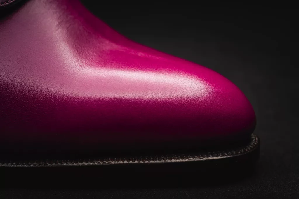 Close-up of the vibrant magenta almond shaped toe of a polished calf leather shoe against a dark background. The shoe has a hand sewn welt and hand stitched oak bark tanned sole.