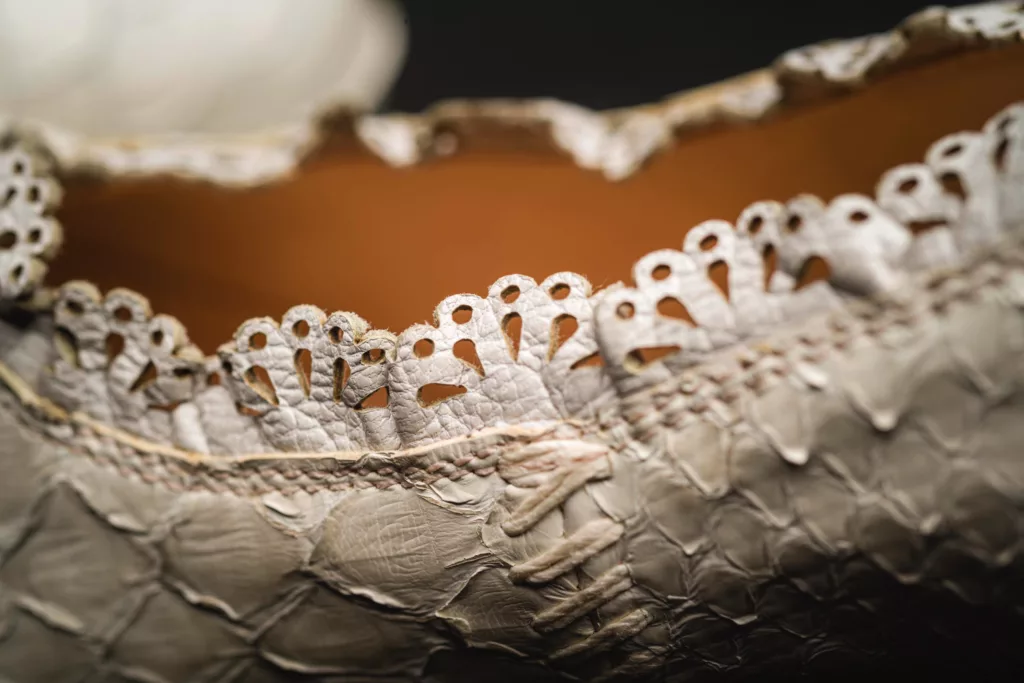 Close-up view highlighting specific elements of a chalky white python skin shoe. The scutes, or scales of the python skin add visual interest along with a decorative leather scalloped edge with fan shaped cut outs and white linen thread hand stitched details at the side seams.