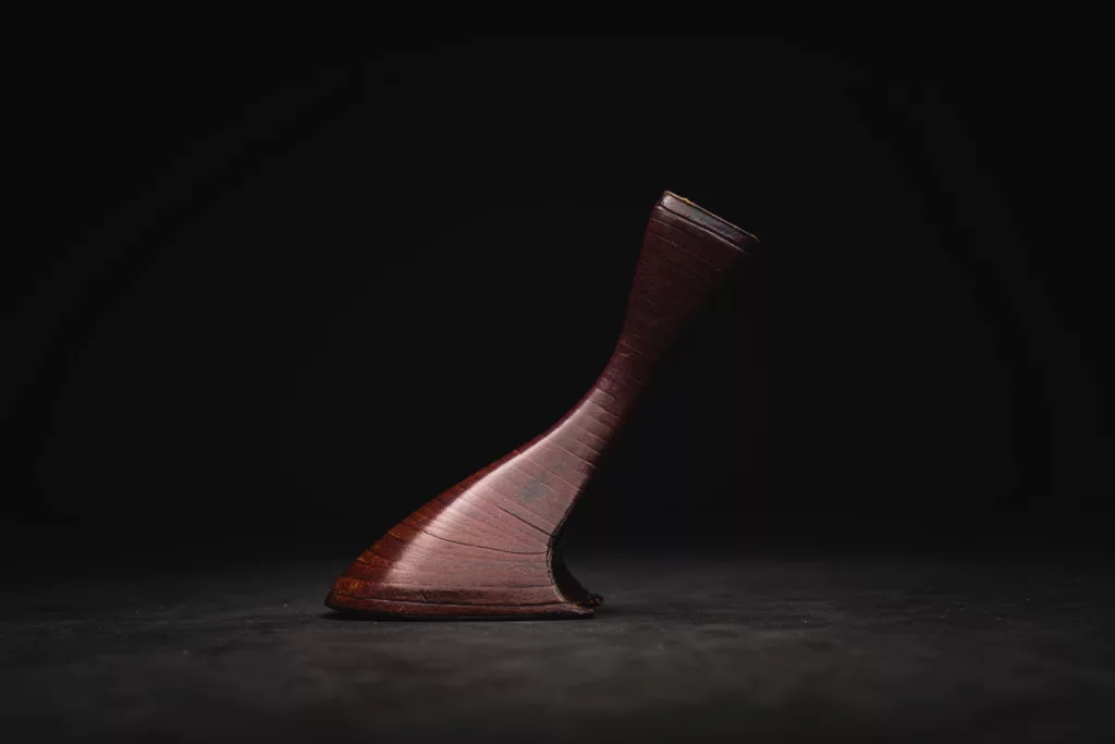 A sculptural single high heel sits on a flat surface against a black background. Formed of individually stacked pieces of oak bark tanned leather stacked one on top of the other, shaped, filed, scraped and polished to create elegant curves and a sumptuously deep rich natural finish.