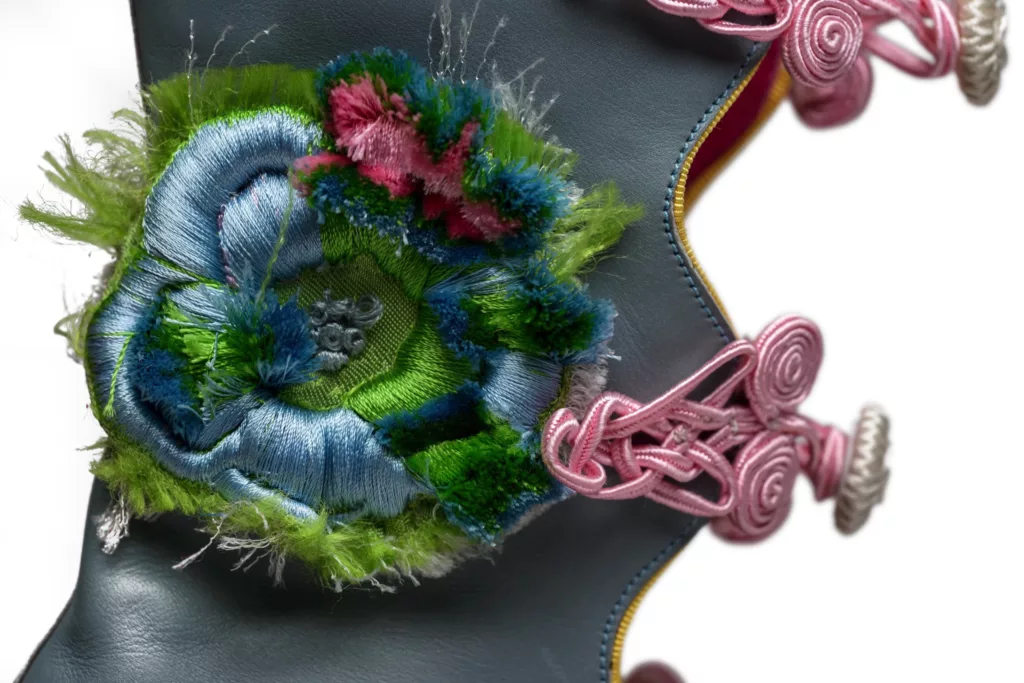 Here you can see a highly detailed part of the Exploding Flowers shoe by Caroline Groves. An open toe, open fronted boot with frogging closures. In pale blue baby calf with covered platforms and heels of vintage French silk in mauve. The ‘Exploding Flowers’ are worked in velvet embroidery of an array of pastel colours. Here blue, green and pink are the main colours on show and a yellow grosgrain beading edge is in sight.