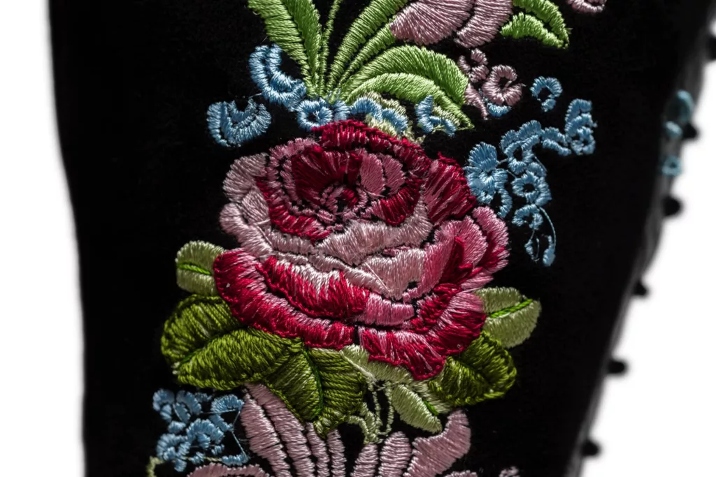 Long leg lace up boot in black suede. This pair has floral silk embroidered leg panels, in pinks, greens and blue grey. Here is a close up that includes red silks in the embroidery of a rose like flower shape.