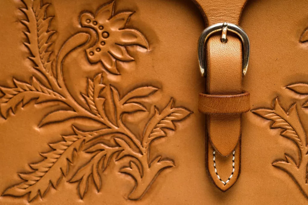 Close up photo of the Valerie oak bark tanned shoulder case bag. The fine detail of oak leaf inspired tooling onto the leather, creating shadows and depth to the piece. Featuring a fastening of brass and the detail of hand stitched linen thread