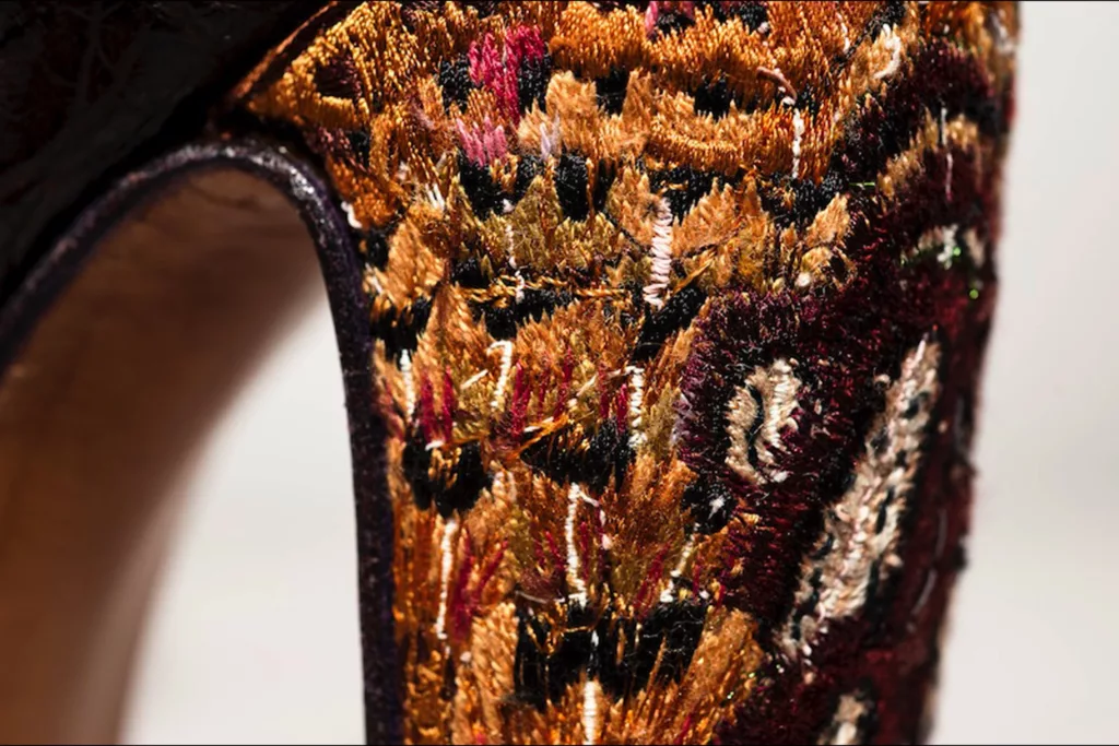 Close-up of a high-heel shoe with intricate embroidery. The embroidery design features a mix of gold, red, black, and beige silk thread, creating a pheasant feather pattern. The stitching is dense and detailed, highlighting the craftsmanship. The image focuses on the heel area, showing the textures of the heel cover.