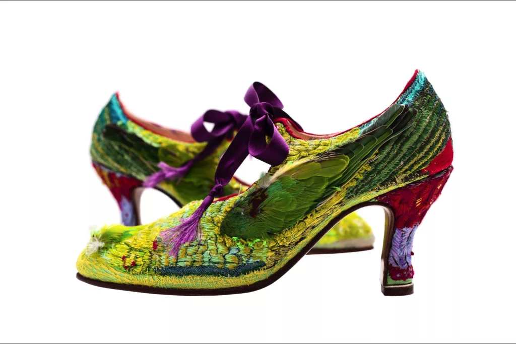 A pair of vibrantly coloured medium Louis heeled shoes decorated with an array of feathers and embroidery stitches including hundreds of French knots. The shoes feature a predominantly green and red pattern, accented with splashes of purple, pink, and blue. A purple satin ribbon ties at the instep.