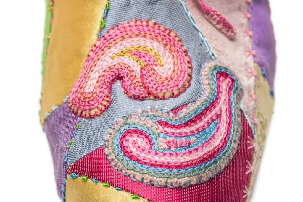 A close up photograph showing the fine details of a court shoe with a single tie on instep. In vintage silks as patchwork and silk embroidery details throughout in soft pastel colours. Here you can see rows of candy colour hand stitched embroidery, a feast for the eyes.