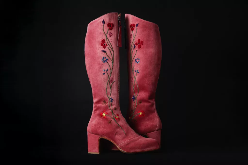 A pair of tall, pink suede boots with a central front zip on each. The hand embroidered boots feature a floral theme in blue, red, and green silk threads showcasing various field flowers and stems. They have a chunky heel.