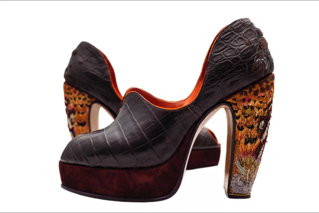 A pair of bespoke high-heeled shoes with a crocodile leather in dark brown. The heels are intricately worked with vibrant yellow, red, and orange pheasant feather embroidery .The shoes feature a deep burgundy suede platform and an orange lining. The shoe is highlighted with an orange grosgrain edge detailing and a heavily worked embroidery covered heel. The sturdy heel’s embroidery is influenced by the pheasant and its distinct feathers. The platform is covered in a deep burgundy suede adding to the tactility of this wonderful creation.