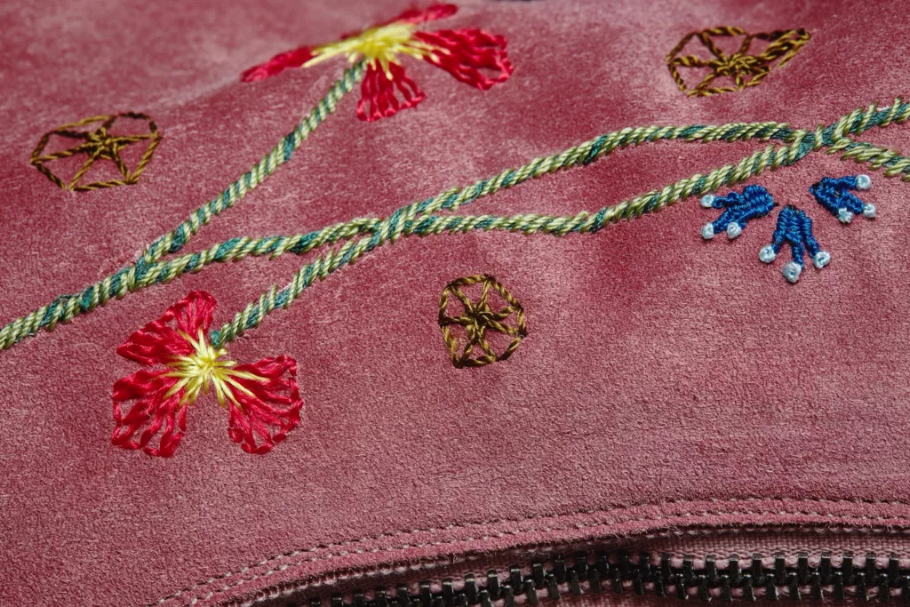 Close-up of intricate floral embroidery on dusky pink baby calf suede. The hand embroidery worked in silk thread features stylised yellow, red, blue & white flowers, green stems intertwined with rope-like strands.