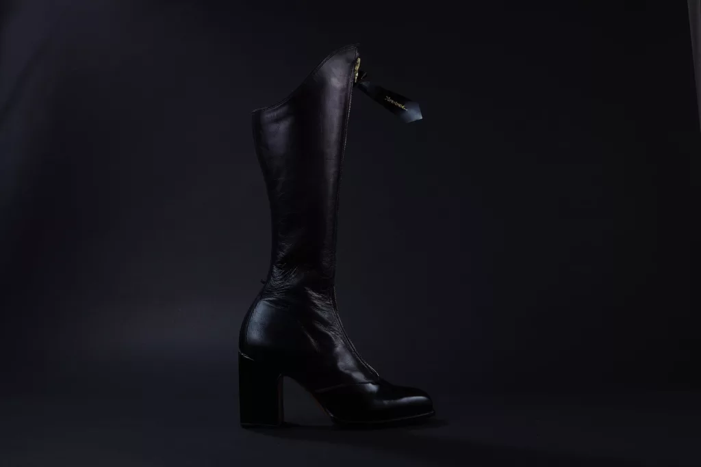 Side profile of a striking black fitted boot. Long leg, zipped front and on a solid square heel. The leather glistening beautifully in the photograph. Metal zip has an oversized leather puller featuring the name of the wearer in Metallic Emboss