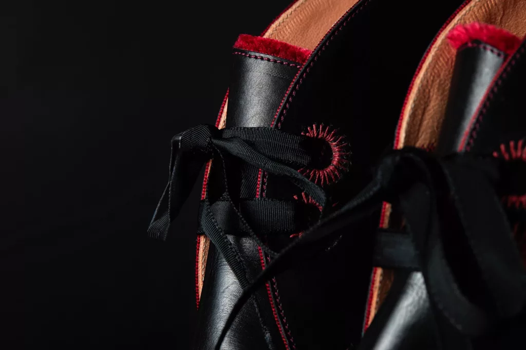 Close up of the eyelets of a black calf leather bootie. Oversized, hand embroidered eyelets of red circular embroidery. Red beading along edges and red velvet embroidery at edge of tongues. All set against fine polished black leather and black grosgrain lacing.