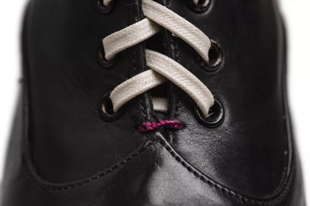 Close-up of a black leather shoe featuring white laces threaded through black eyelets. A small purple stitching detail is visible near the bottom lace. The leather has a smooth, glossy finish, and the stitching and lacing are in focus, emphasizing the shoe's craftsmanship.