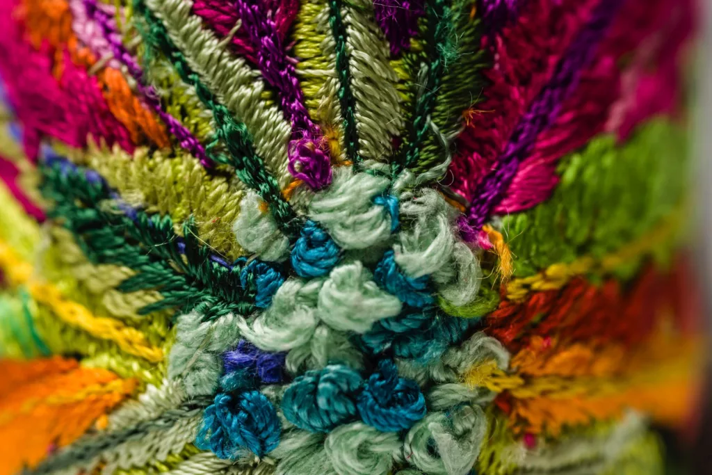 Close-up image of colorful, intricate embroidery showcasing a variety of vibrant threads. The design features a mix of greens, blues, pinks, oranges, and purples with detailed patterns and textures, creating the appearance of floral and leaf motifs. The stitches are densely packed and richly textured.