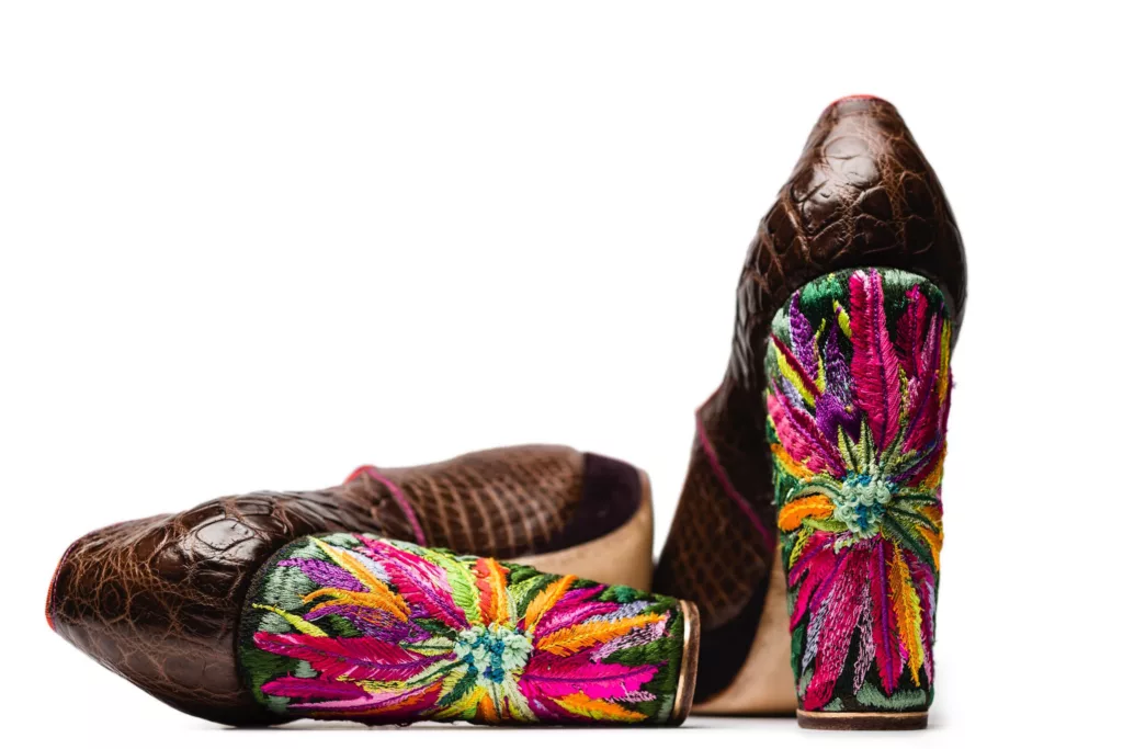 A close-up image of two high-heeled shoes with a brown crocodile-skin pattern. The chunky heels are adorned with vibrant embroidered floral designs featuring bright pink, orange, yellow, purple, and green threads, creating a striking contrast against the dark leather. The background is plain white.