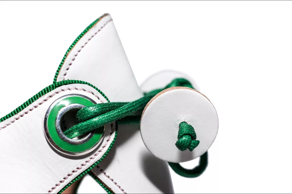 Close-up of a white leather shoe being laced with green laces. The laces are threaded through a metal eyelet with a green interior and tied in a knot, secured by a white leather disk with a green edge. The background is white, drawing attention to the detailed lacing and materials used.